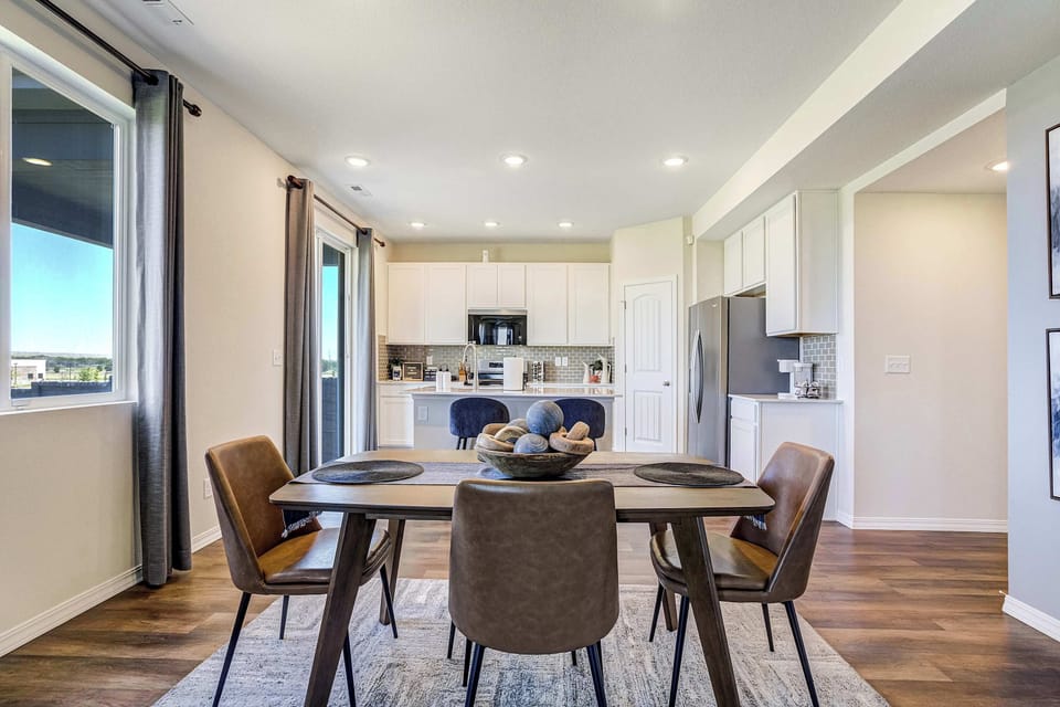 The dining space flows seamlessly into the kitchen, with a wood table and upholstered chairs set beneath a row of windows. White cabinetry and stainless appliances are visible just beyond, tying the main floor together.