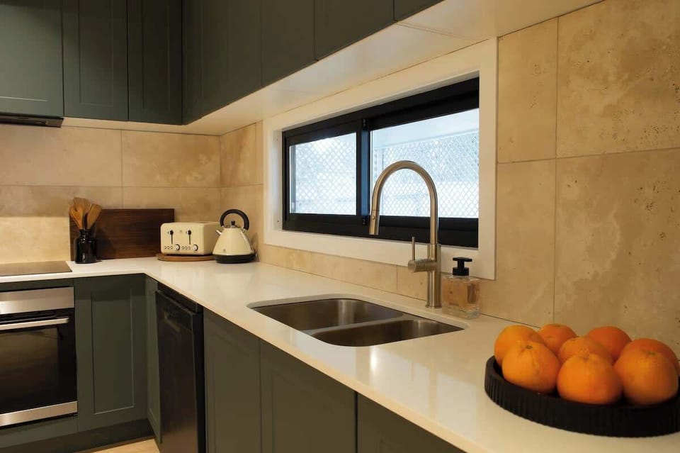 Enjoy cooking in this stylish kitchen, featuring dark olive green cabinets, smooth white countertops, an undermount double-basin sink, and natural tile backsplash.