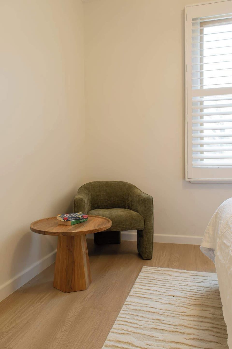Unwind in this cozy reading nook, featuring a olive green barrel armchair, a round wooden side table, a textured area rug, and window shutters that let in soft natural light.