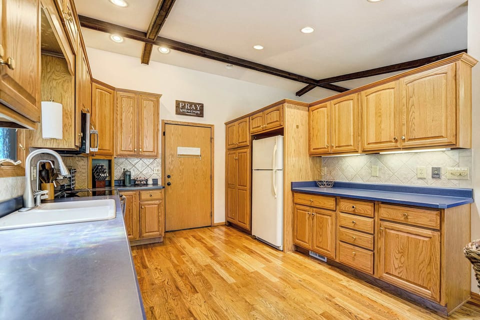 This angle reveals the full galley-style layout, with a white refrigerator and an additional built-in wall oven anchoring one end. Exposed wood beams stretch overhead, while warm hardwood flooring runs the length of the kitchen.

