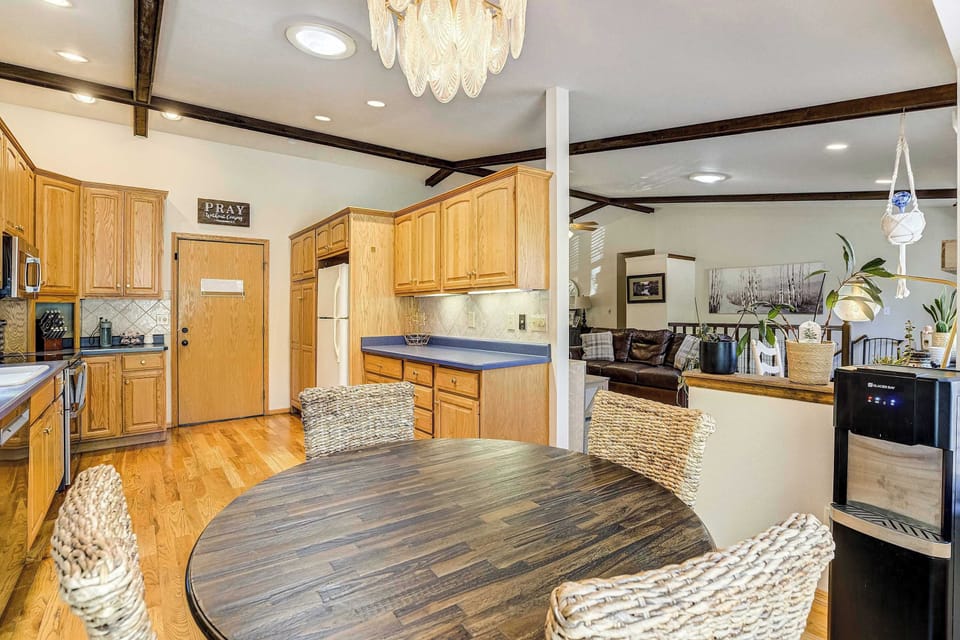 The kitchen centers around a round wood table with woven accent chairs, set beneath a crystal chandelier. Warm oak cabinetry, stainless appliances, and dark countertops carry through, all resting on rich hardwood flooring.

