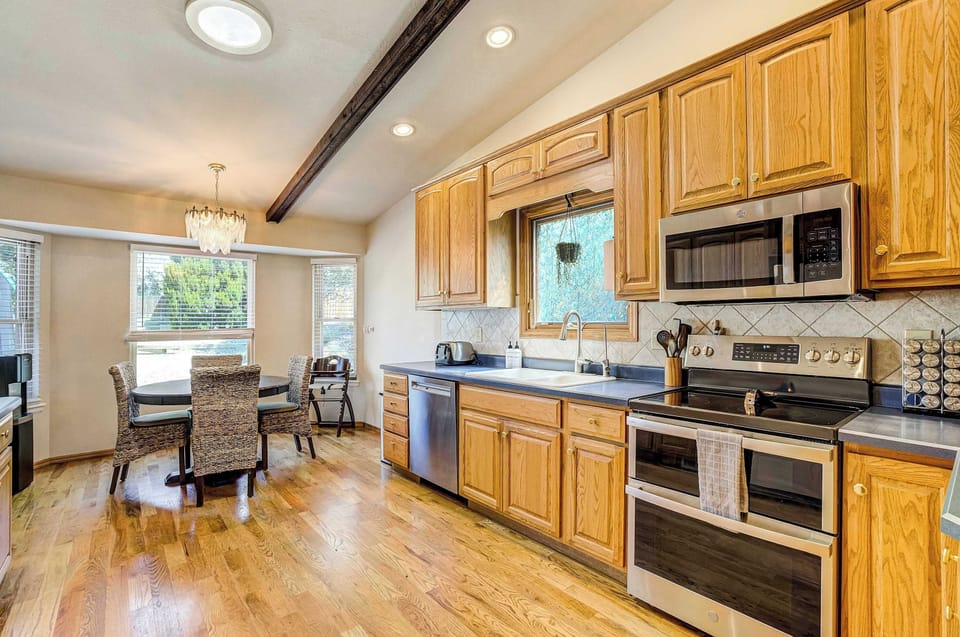 From a wider angle, the fully stocked kitchen shows off oak cabinetry, a tile backsplash, and stainless steel appliances beneath an exposed wood beam ceiling. A small four-seat table offers a casual spot for quick meals.

