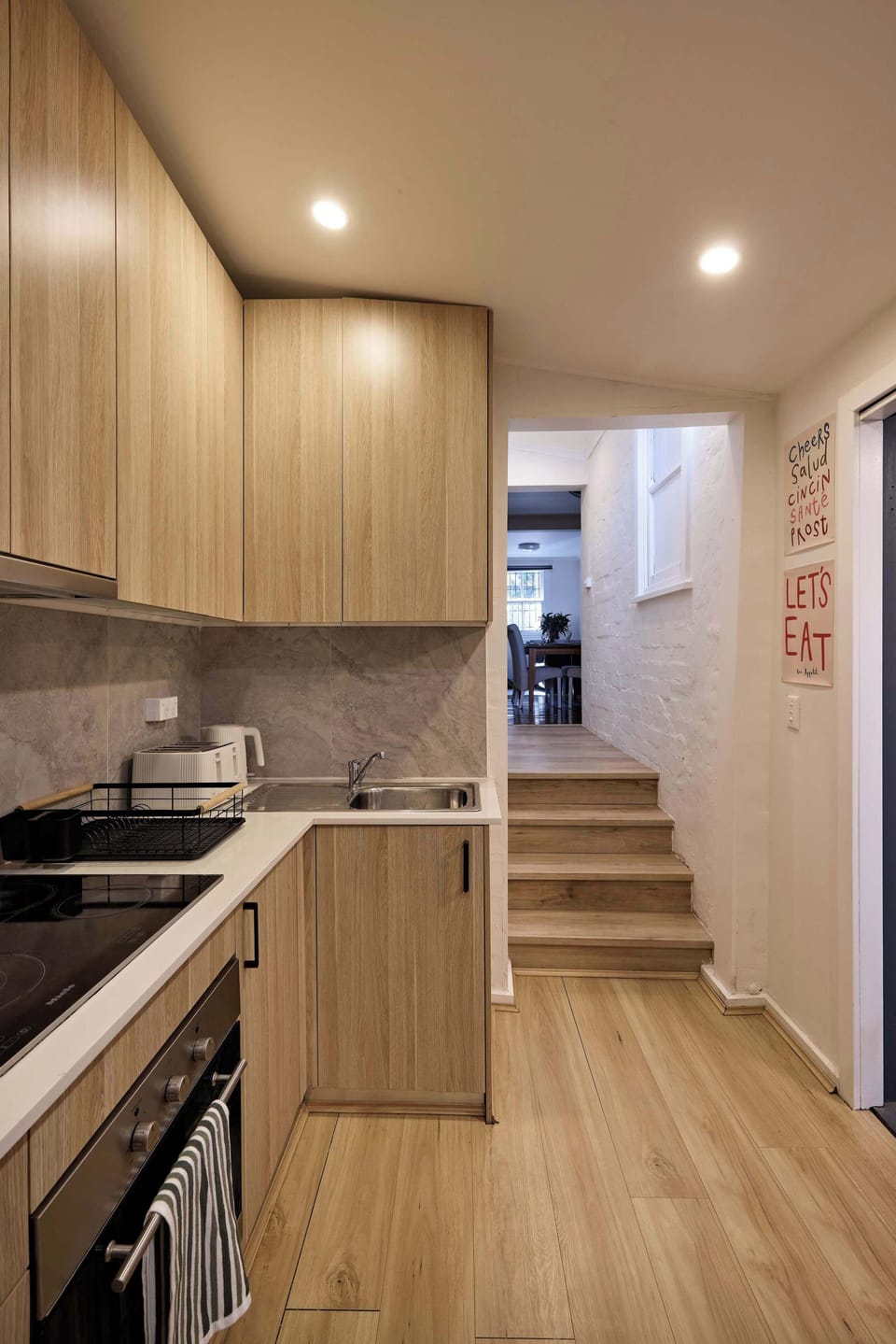 Prepare home-cooked meals in this modern kitchen, equipped with warm light wood cabinetry, an integrated cooktop and oven, sleek countertops, and steps leading into the adjoining living spaces.