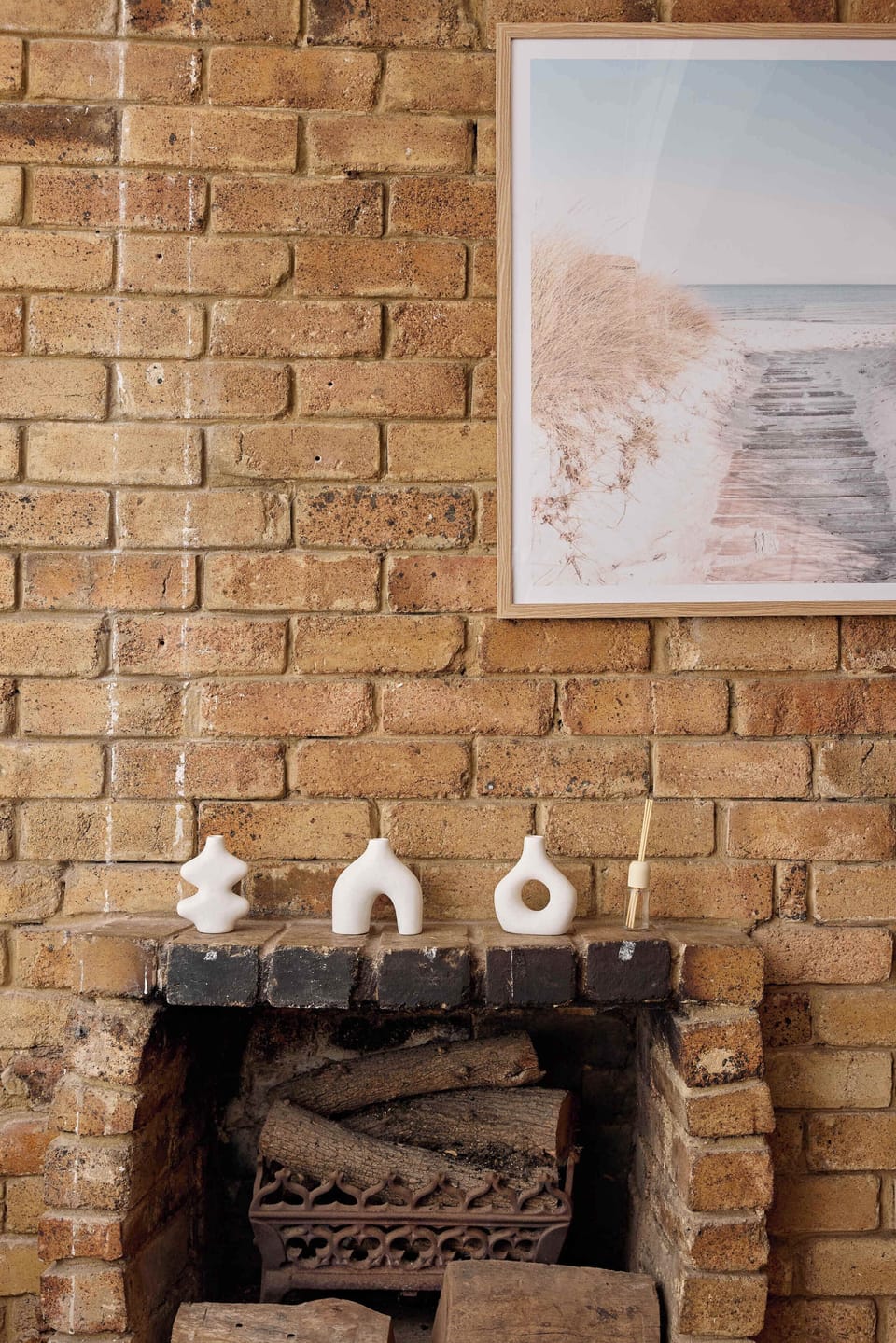 Admirers of rustic details will appreciate this cozy brick fireplace alcove, featuring stacked firewood, decorative ceramic accents along the ledge, and a soothing framed art print above.