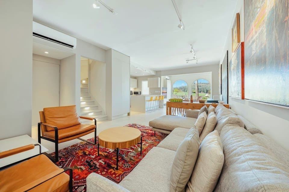 Angle of the main living space showcasing the gray sectional sofa, leather accent chairs, a round coffee table on a vibrant oriental-style rug, and a sleek modern staircase with step lighting.