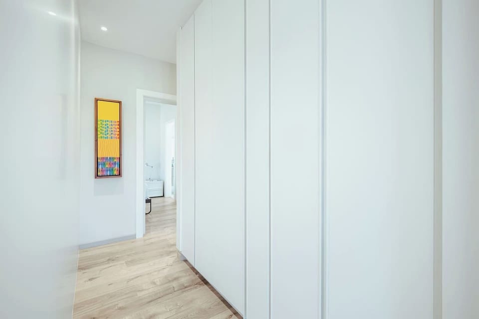 Clean, modern hallway featuring seamless white floor-to-ceiling closet storage, light wood flooring, and a colorful vertical art piece.