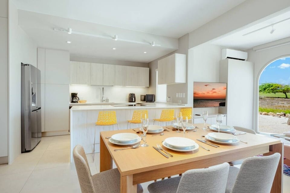 Bright kitchen and dining space featuring a breakfast bar with vibrant yellow stools, set dining table for six, stainless steel appliances, and large windows.