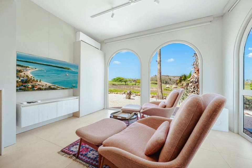 Chic, sunlit lounge space featuring two dusty-rose lounge chairs with matching ottomans, a large wall-mounted flat-screen TV, and sweeping views of tropical palm trees through arched windows.