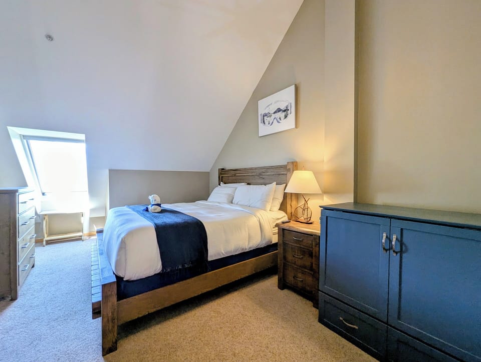 Step into your peaceful retreat under the sloped ceiling, where soft light filters through the skylight and crisp linens invite restful sleep on a beautifully crafted wooden bed.