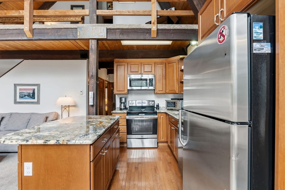 Your open-concept kitchen invites you to cook and gather with modern stainless-steel appliances, granite counters, and warm wood cabinetry under soaring timber beams.