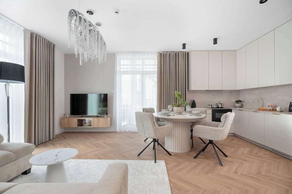 A wide-angle shot shows the living room with a TV console, beige sofa, and round dining table. Off-white kitchen cabinets line the far wall.