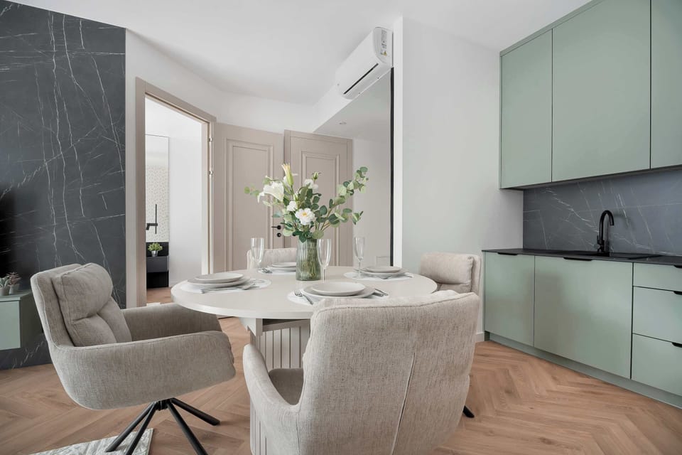 A perspective from behind textured dining chairs showcases a round table set for a meal. Sage-green kitchen cabinets line the background.
