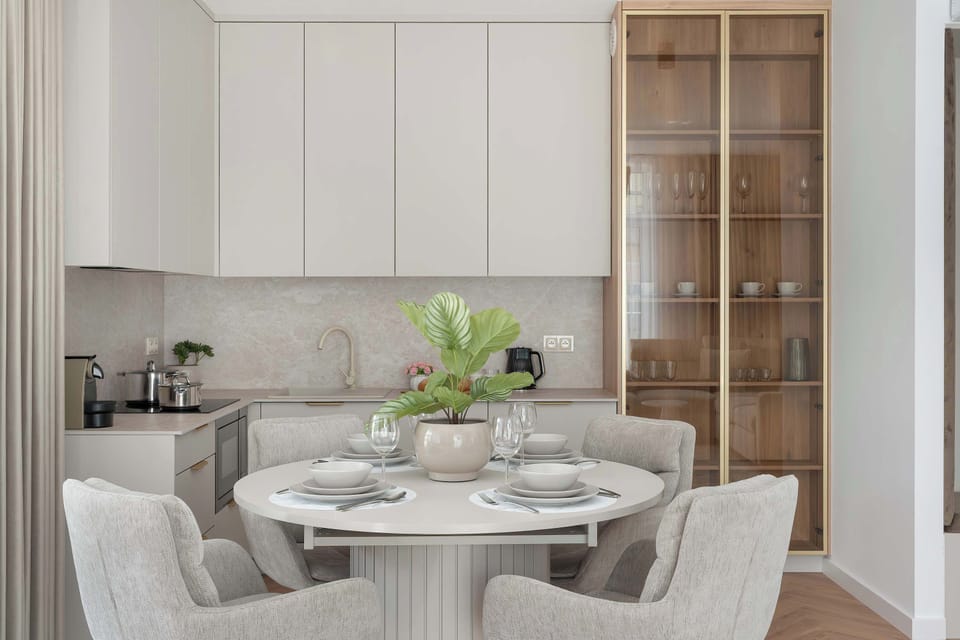 A dining setting for four includes a round table and upholstered chairs next to off-white cabinetry. A tall glass display unit sits on the right.