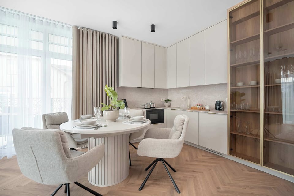 A round fluted dining table with beige swivel chairs sits near off-white kitchen cabinets. A glass display cabinet frames the side of the dining nook.