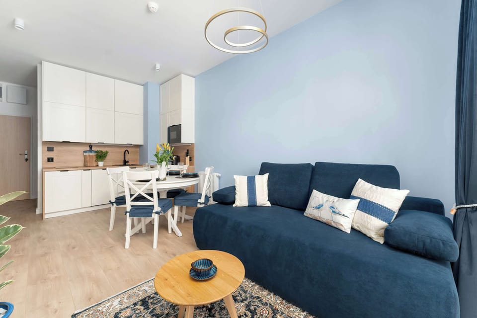 Looking across the entire open-plan space unifies the kitchen, dining, and living areas. White cabinets, a white dining set, and the blue sofa create a light, cohesive design. Wood flooring extends throughout.
