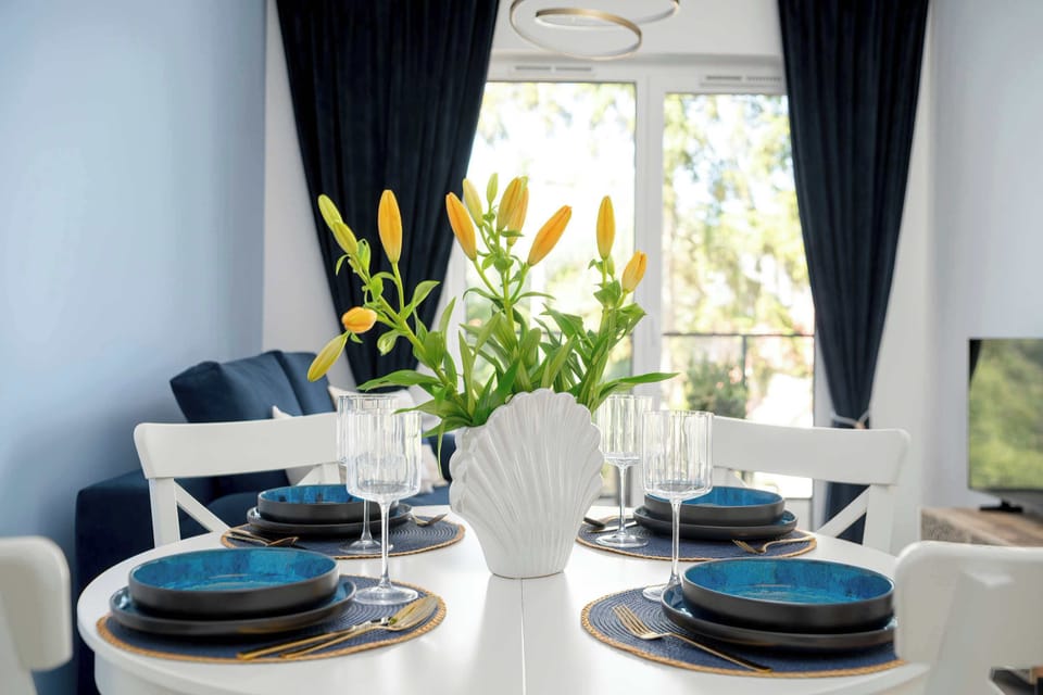 A closer look at the dining table shows it set with blue plates and glassware. A vase of yellow flowers provides a centerpiece. Through the window, green treetops are visible.