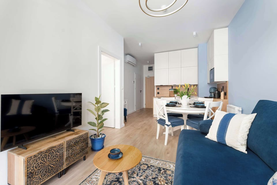 An open-plan area shows the dining table with chairs next to the living room setup. A modern light fixture hangs over the table on the blue-painted wall. Wicker details on the TV stand add natural texture.