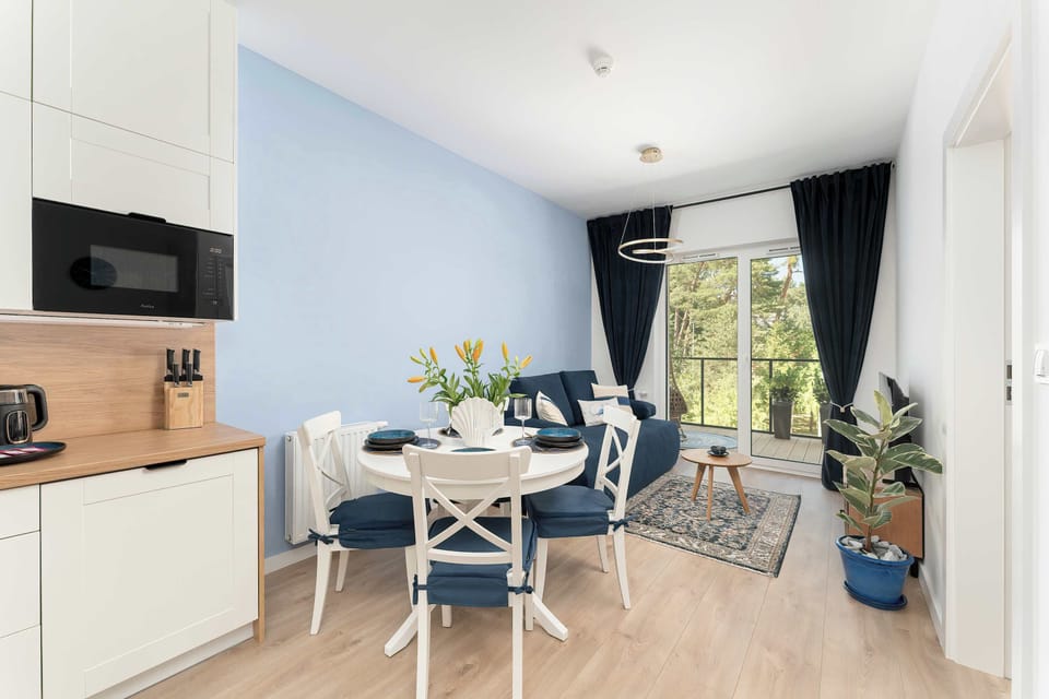 A wide shot shows the seamless connection between the modern white kitchenette and the dining space. Sunlight streams from the adjacent living room area. Potted plants add life to the clean environment.