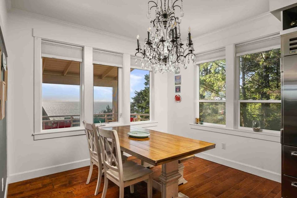 Sunlit dining space featuring large windows framed by ocean and forest scenery.