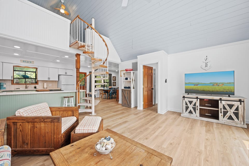 Unwind in your bright coastal living space featuring vaulted ceilings, warm wood tones, and a spiral staircase leading to your private loft. The open layout connects you to a full kitchen, making relaxation effortless.