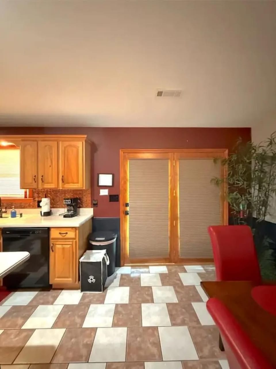 Kitchen area with natural wood cabinetry, black dishwasher, checkered tan and white tile floor, and red dining chairs.