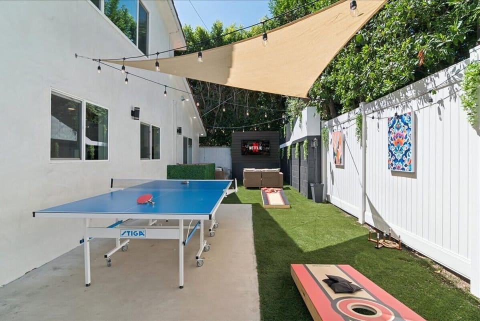 Backyard during the day with outdoor aluminum Stiga ping-pong table, corn hole, and large TV with sofa area for hanging out