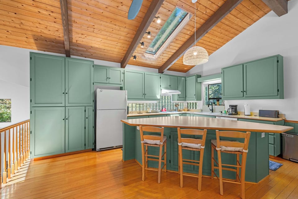 Pull up a barstool at the angled kitchen island—sage cabinetry, a woven pendant, and a skylight overhead keep mornings bright.