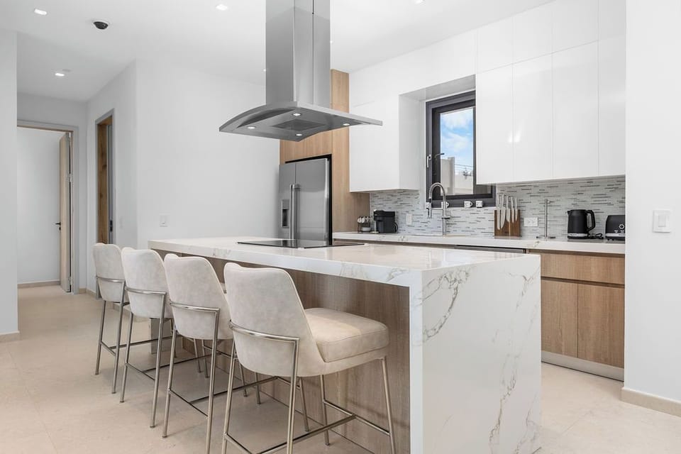 A hub for connection, not just cooking. The waterfall-edge island with cream barstools is perfect for morning coffee, casual meals, or wine with loved ones — all set against bright, airy white tile and walls that make you feel right at home.
