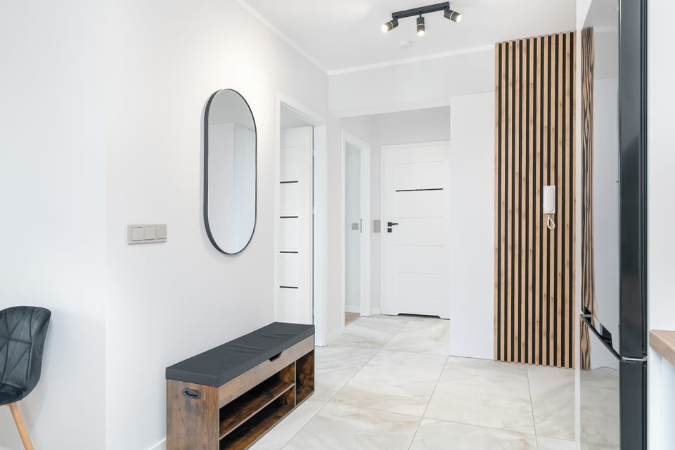 A clean entrance hallway with smooth white walls, an oval mirror, and a small bench. Vertical wooden slats add a contemporary architectural touch.