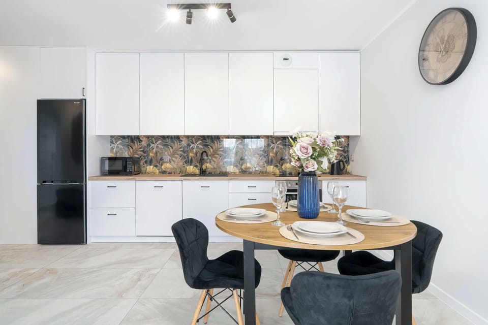 A bright kitchen and dining space featuring white upper cabinets, a black refrigerator, and a dark dining table with chairs. Light flooring enhances the open feeling.