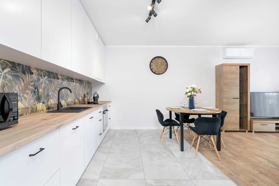 A wide perspective of a bright modern kitchen with white cabinetry and wooden accents. Light floor tiles blend seamlessly into wood-style flooring in the living space.