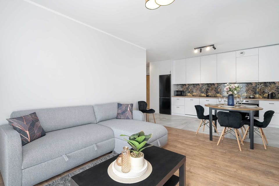 An open-plan living room showing a light L-shaped patterned sofa and a sleek coffee table. The modern white kitchen and dining space are visible in the background.