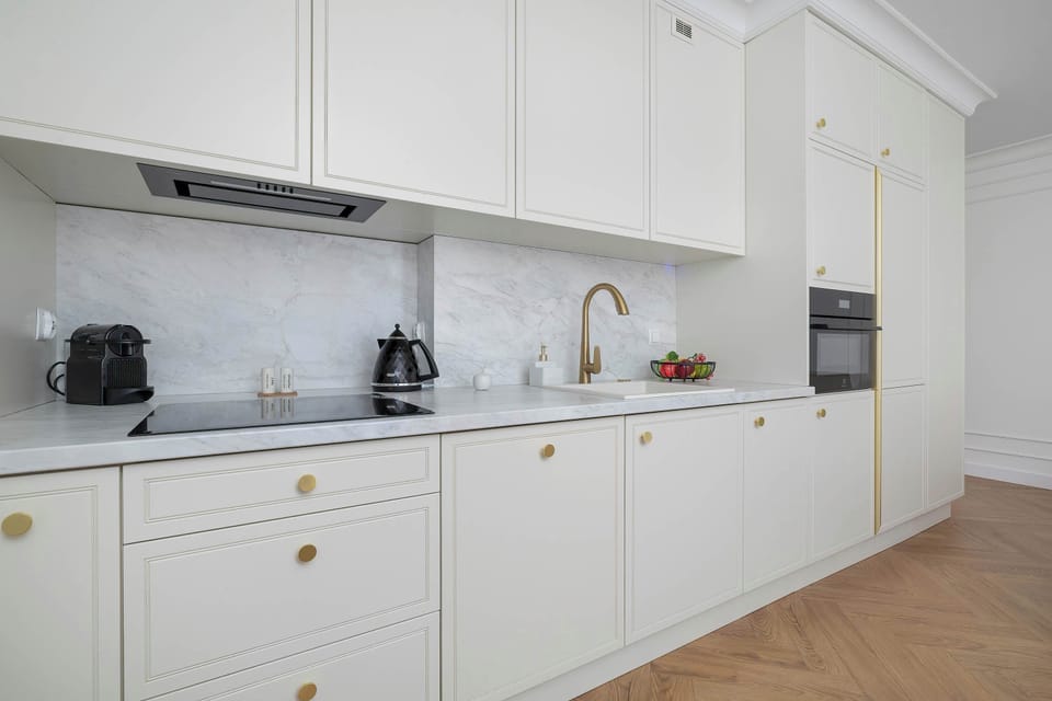 A broad view of a long kitchen counter equipped with an induction cooktop, sink, and built-in appliances. White paneled cabinetry complements the light wood flooring.