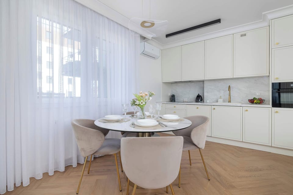 A bright dining space featuring a round table with dinnerware, adjacent to a clean white kitchen. Large windows let in plenty of daylight.
