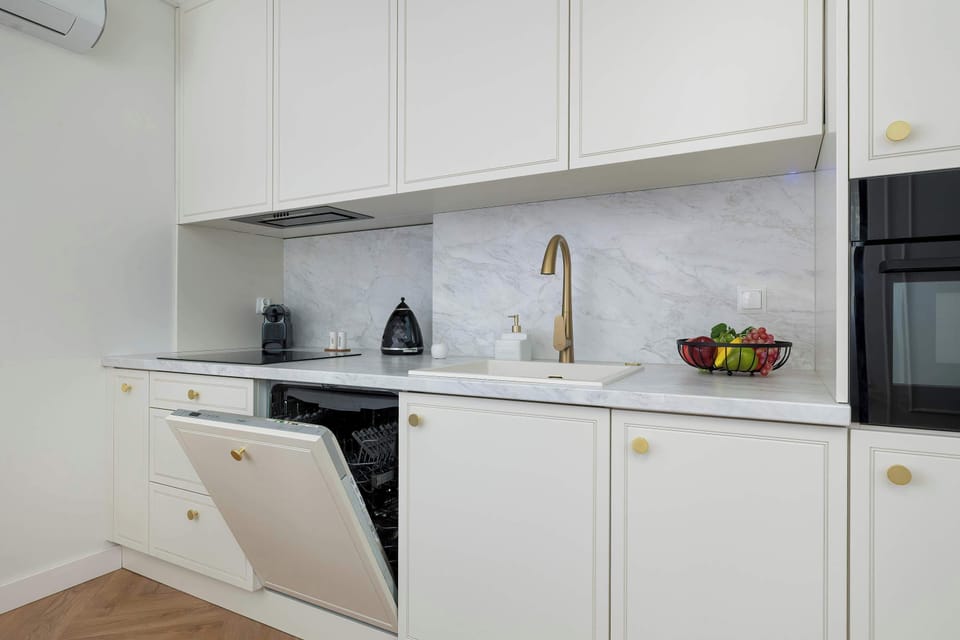 A close-up shot of a modern kitchenette showing an open dishwasher integrated into white cabinetry. Marble splashback and gold finishes highlight the upscale design.