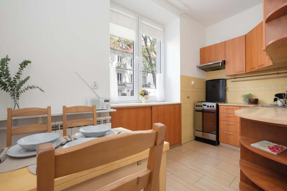 The kitchen view highlights the adjacent dining arrangement, featuring a sturdy wooden table set for a meal. Large windows fill the entire cooking and dining space with natural daylight.