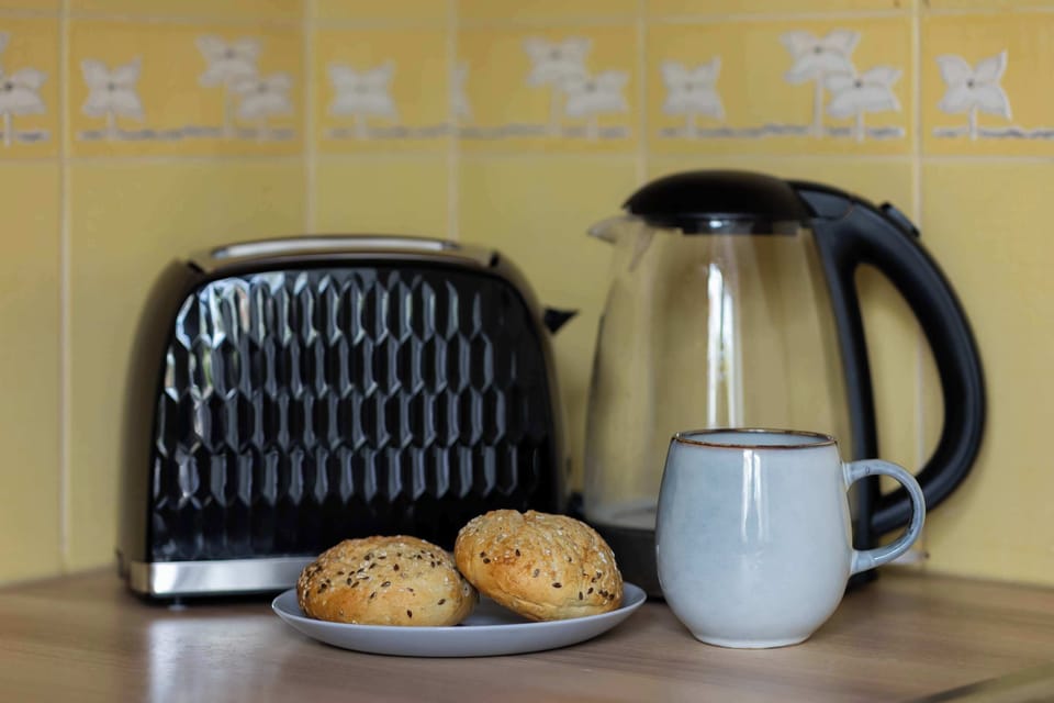 A close-up shot on the kitchen counter focuses on a modern black toaster and a glass electric kettle filled with water. A white ceramic mug and some fresh bread rolls rest nearby on a plate.