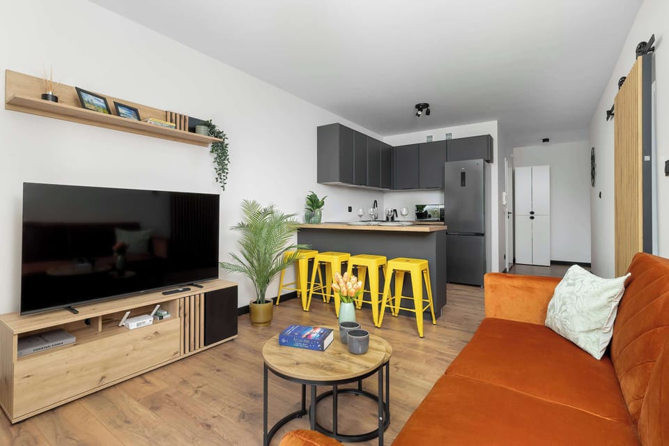 An open-plan view combines the living area and kitchenette zone, showing the back of the media console and a kitchen island with yellow barstools. The seamless layout emphasizes a sense of spaciousness.