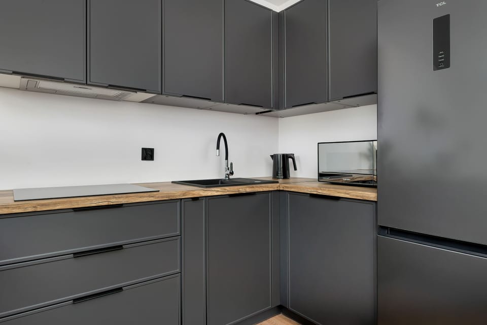 A close-up of the kitchenette counter highlights the combination of dark grey cupboards and a clean wooden surface. Sockets and a black appliance are neatly arranged along the wall.