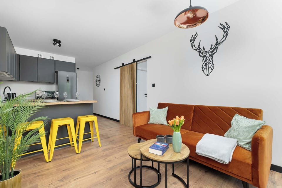 A wide shot of the open living and dining area reveals an island counter paired with bright yellow stools. The sofa sits comfortably along the adjacent wall underneath minimalist line art decoration.