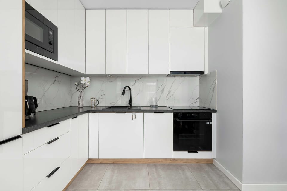 A sleek, minimalist white kitchenette features clean cabinetry, dark countertops, and integrated appliances. The well-organized space looks bright, functional, and ready for meal preparation.