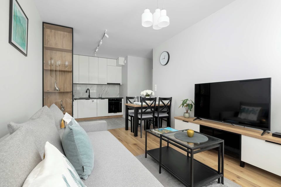 Looking across the living room reveals a view towards the dining table and the adjoining white kitchen. A large flat-screen television is mounted above a sleek media console on the right side of the room.