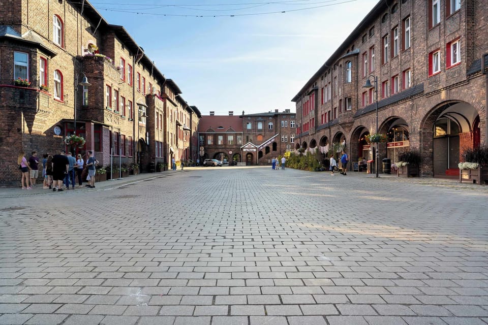 A wide cobblestone plaza stretches between historic red-brick buildings with ground-floor arcades. Sunlight illuminates the open pedestrian street layout.