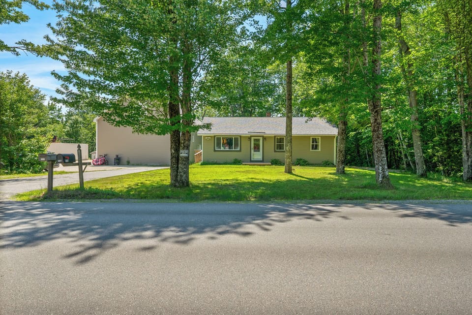 Arrive at this private ranch-style home set back from the paved road in Lamoine, Maine. View highlights the grass lawn, mature shade trees, front entrance, and the spacious driveway entryway leading into the property’s loop parking setup.