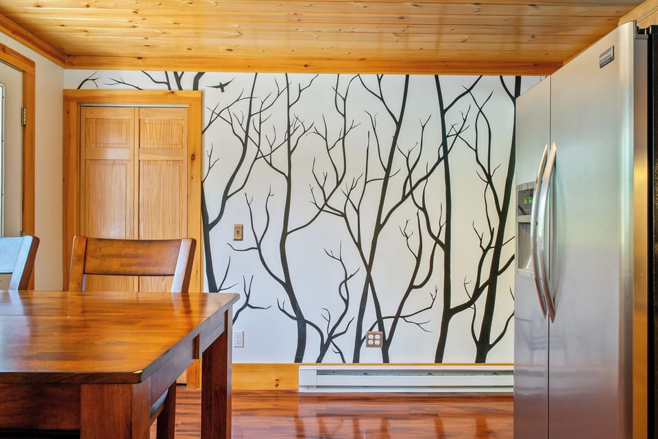 Look across the dining area to reveal the stainless steel side-by-side refrigerator equipped with an exterior ice and water dispenser. The shot highlights the custom tree accent wall, hardwood flooring, and bifold doors to the kitchen pantry.