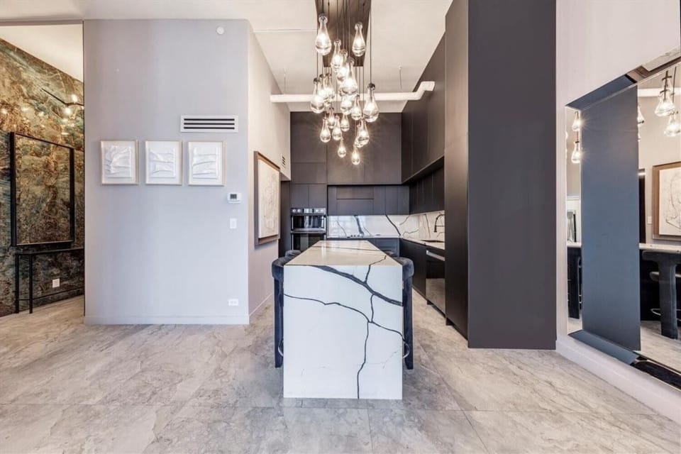 Striking kitchen view highlighting the bold veined marble waterfall island beneath a glamorous cluster of glass pendant lights. Matte black cabinetry and artful stone accents create a sophisticated, high-design culinary space.