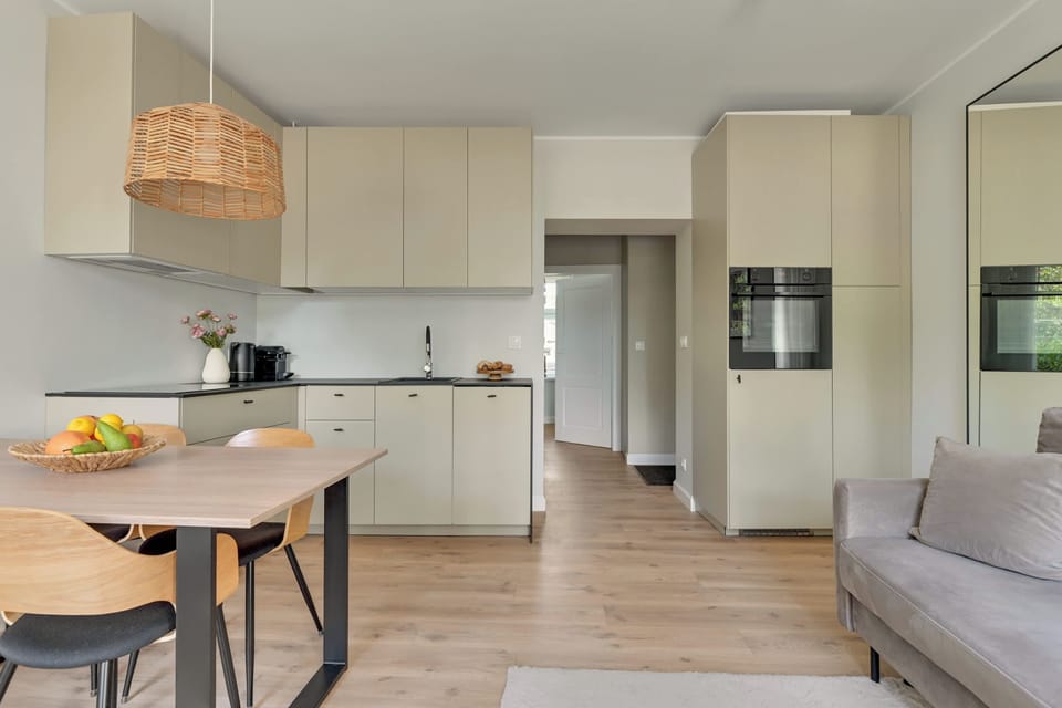 Przestronny i elegancki salon z wygodną sofą, jasną kolorystyką i dużymi oknami zapewniającymi naturalne światło. Spójna aranżacja, drewniane akcenty i przytulna strefa jadalniana tworzą komfortową przestrzeń do wypoczynku.