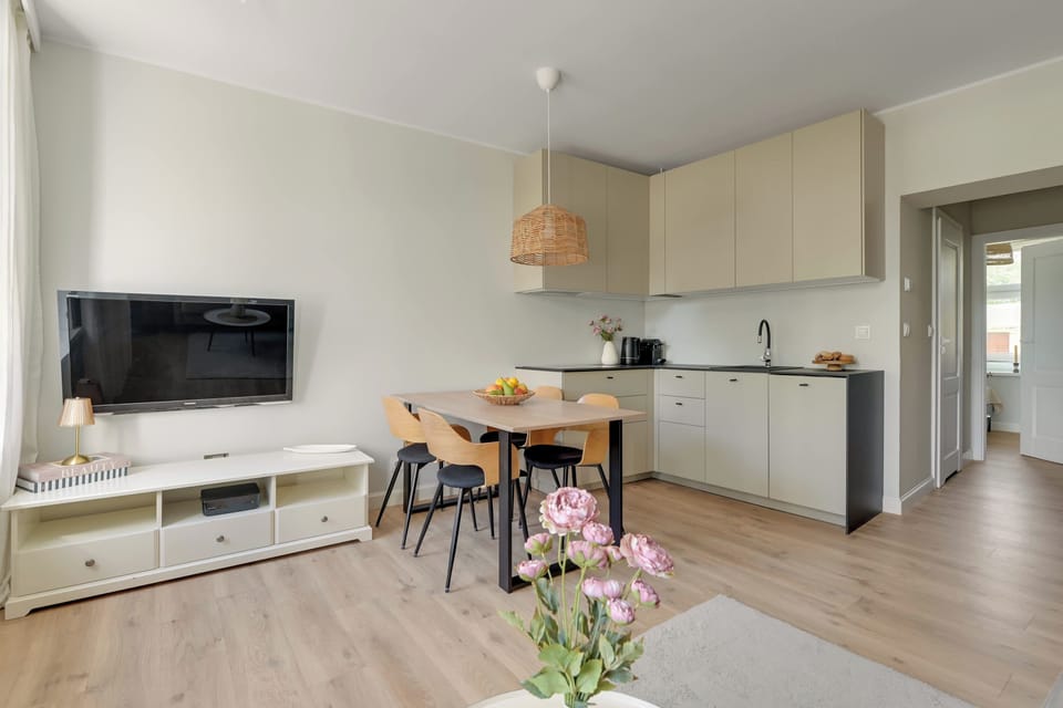 Przestronny i elegancki salon z wygodną sofą, jasną kolorystyką i dużymi oknami zapewniającymi naturalne światło. Spójna aranżacja, drewniane akcenty i przytulna strefa jadalniana tworzą komfortową przestrzeń do wypoczynku.