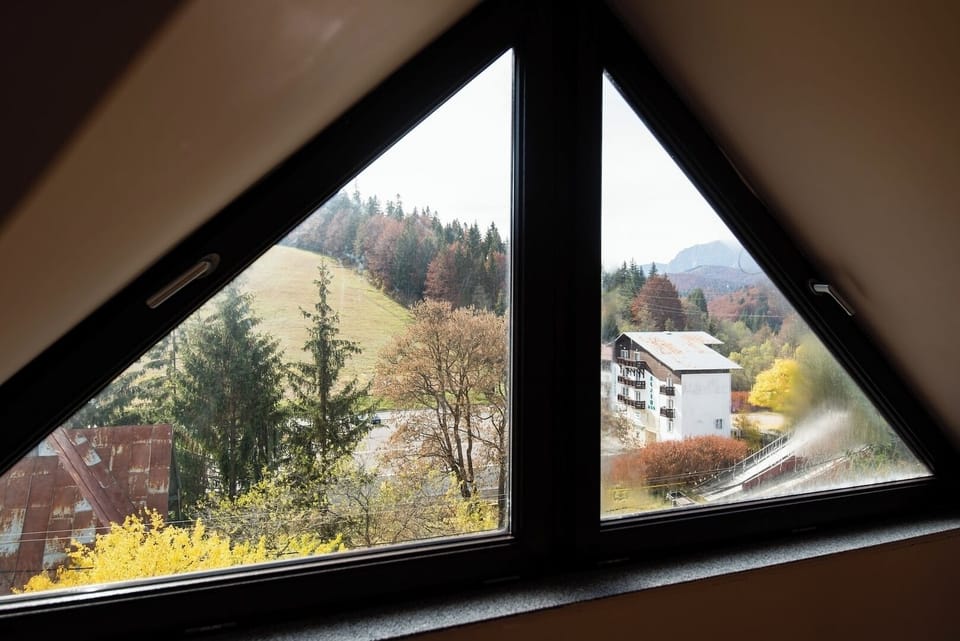 Open mountain views through the living room's triangular window — pine forest, Predeal rooftops and rolling hills in one frame. One of the most distinctive views available in this Predeal apartment.