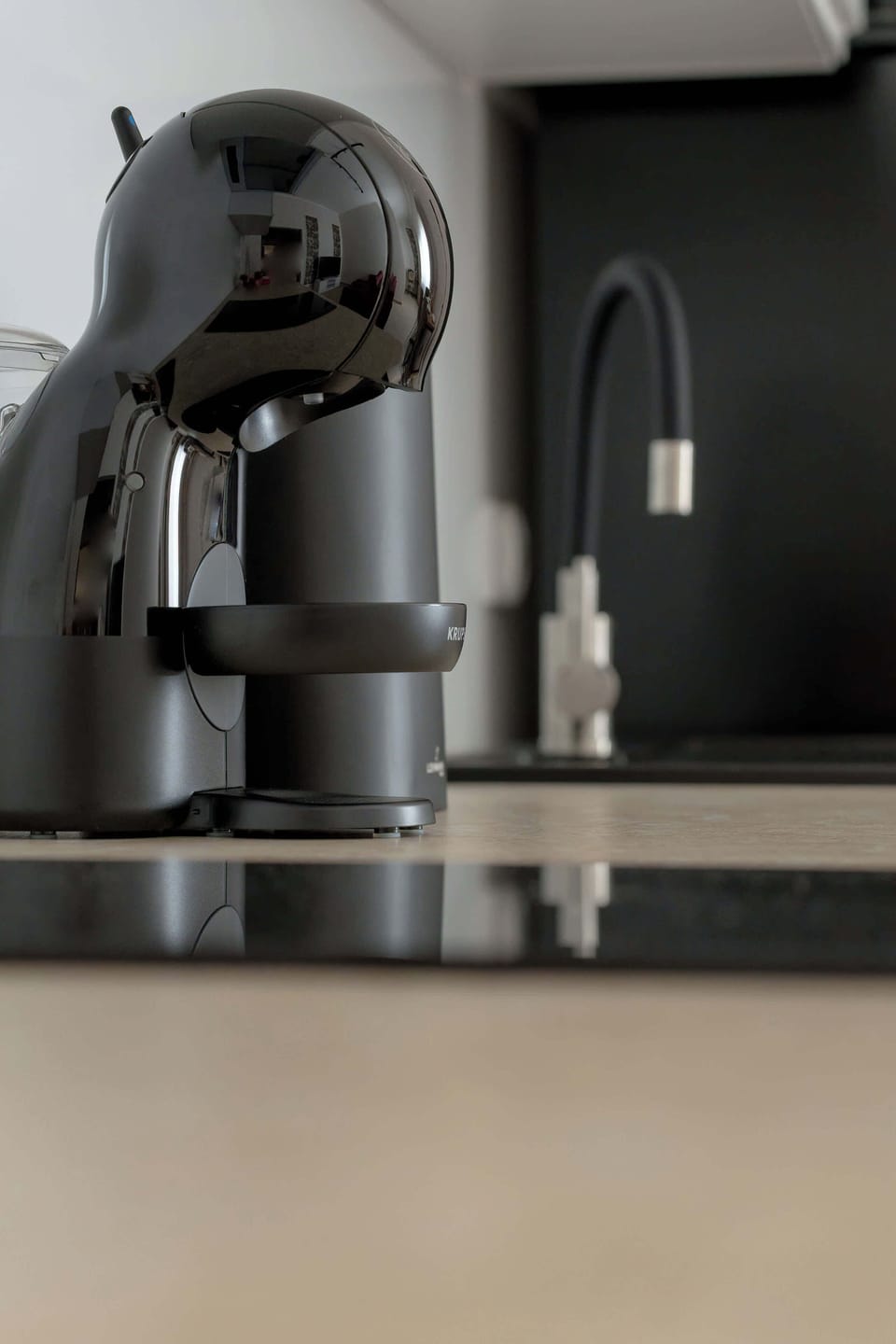 A close-up shot focuses on a black capsule coffee maker resting on a glossy dark countertop. The background shows a clean wall and polished kitchen surfaces.